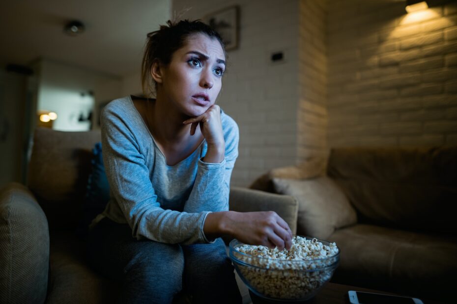 Young woman sitting in the dark and watching TV while eating popcorn at home.